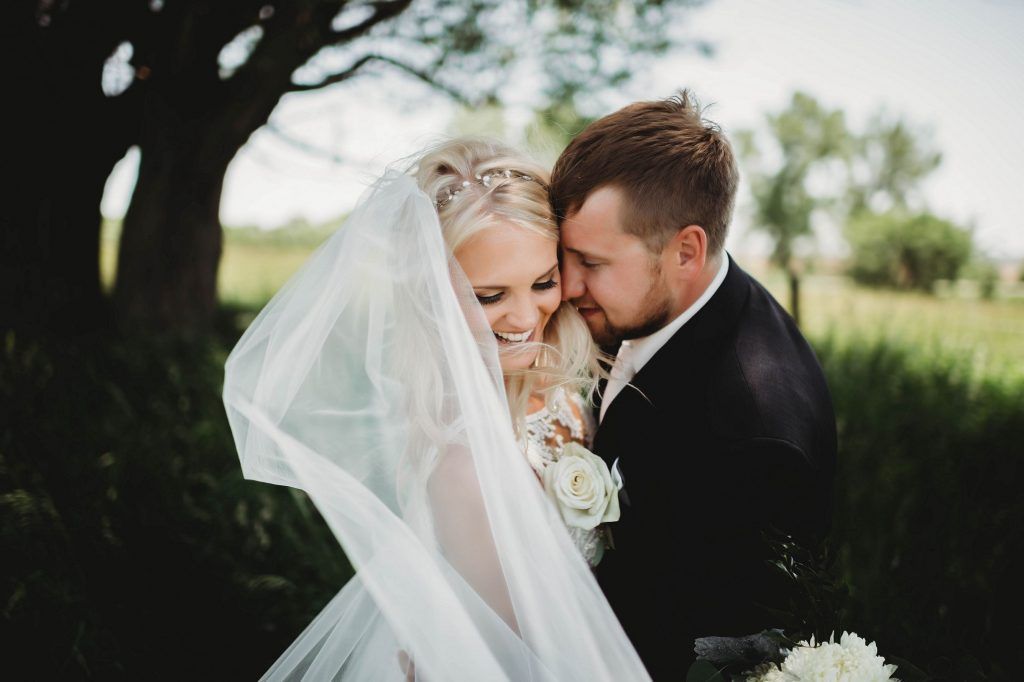 Bride and groom embracing, covered by a veil, outdoor setting with greenery.