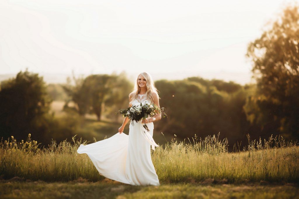 Bride in flowing white dress holding bouquet, standing in a field with trees in the background.