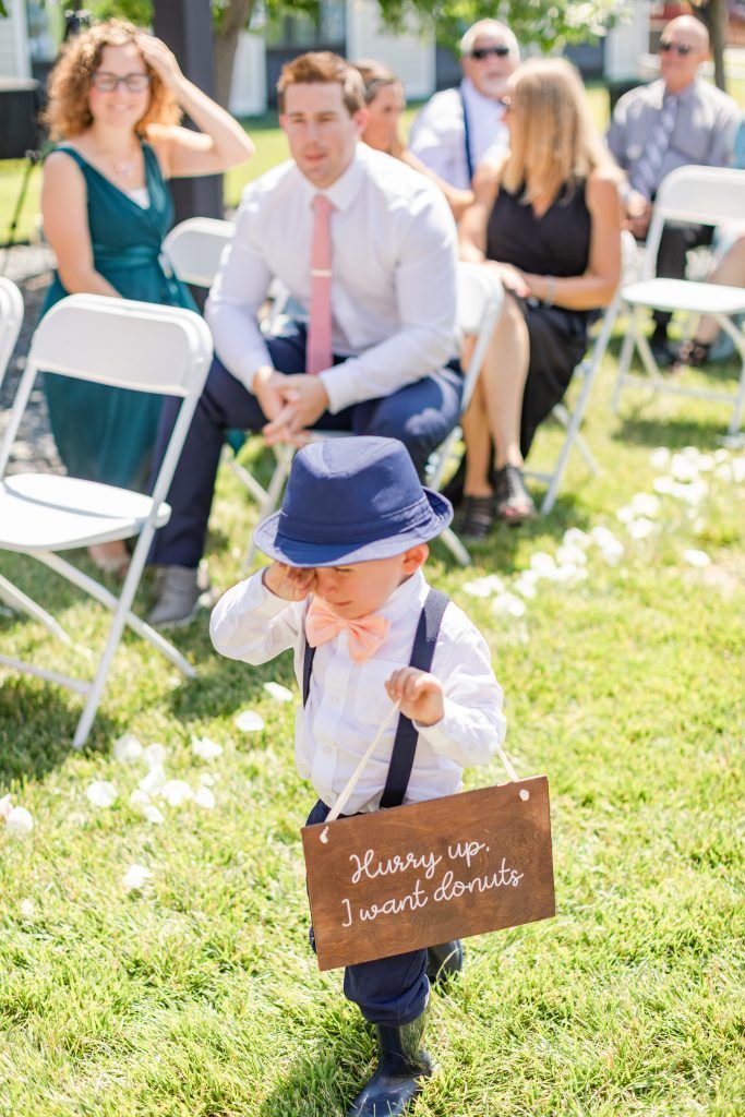 Young child in a blue hat and suspenders, carrying a sign at a wedding ceremony.