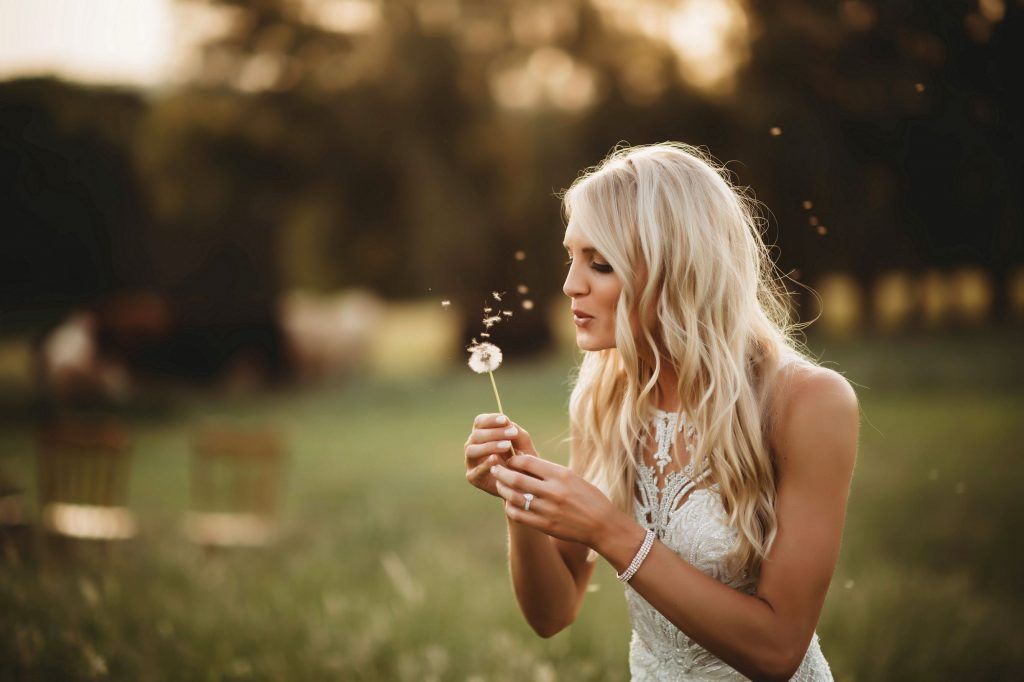 Blonde woman blowing dandelion in a field. Warm sunlight bathes her and the tall grass.