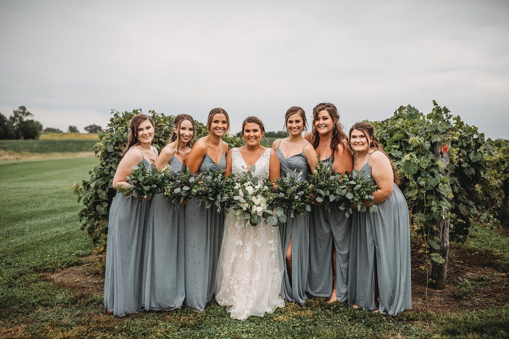 Bride and bridesmaids pose in a vineyard, wearing blue gowns, holding greenery, and smiling.