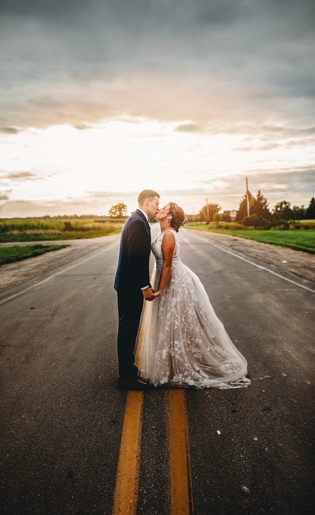 Couple kissing on a road at sunset; woman in wedding dress, man in suit.