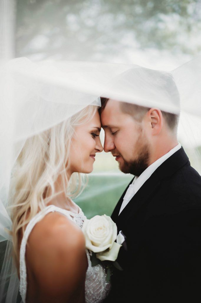 Bride and groom, face to face under a veil, eyes closed. White dress, black suit. Holding a rose.