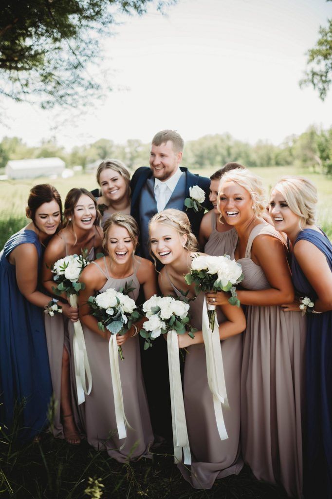 Groom smiles with bridesmaids in a field. Bridesmaids hold bouquets, wearing gray and blue dresses.