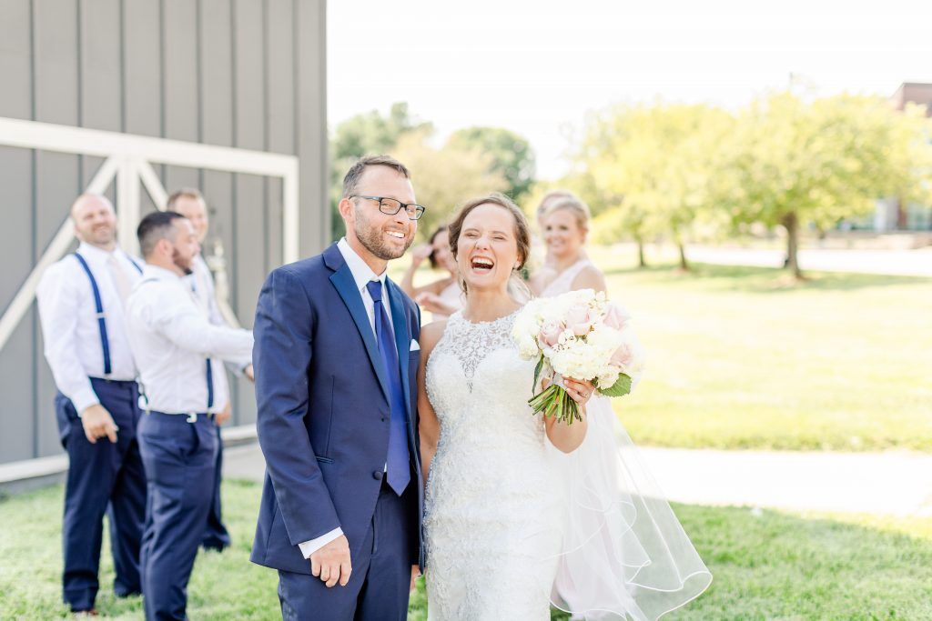 Bride and groom smiling outdoors, wedding party in background near a grey building.