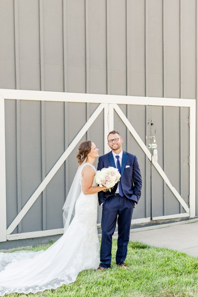 Wedding couple poses in front of a gray barn door. Bride in white dress, groom in blue suit.