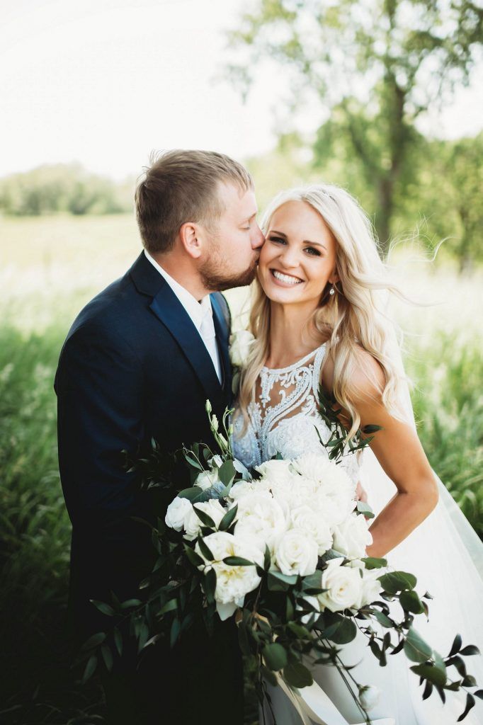 Groom kissing bride's cheek outdoors. Bride holds bouquet; both smile. Green field and trees in background.