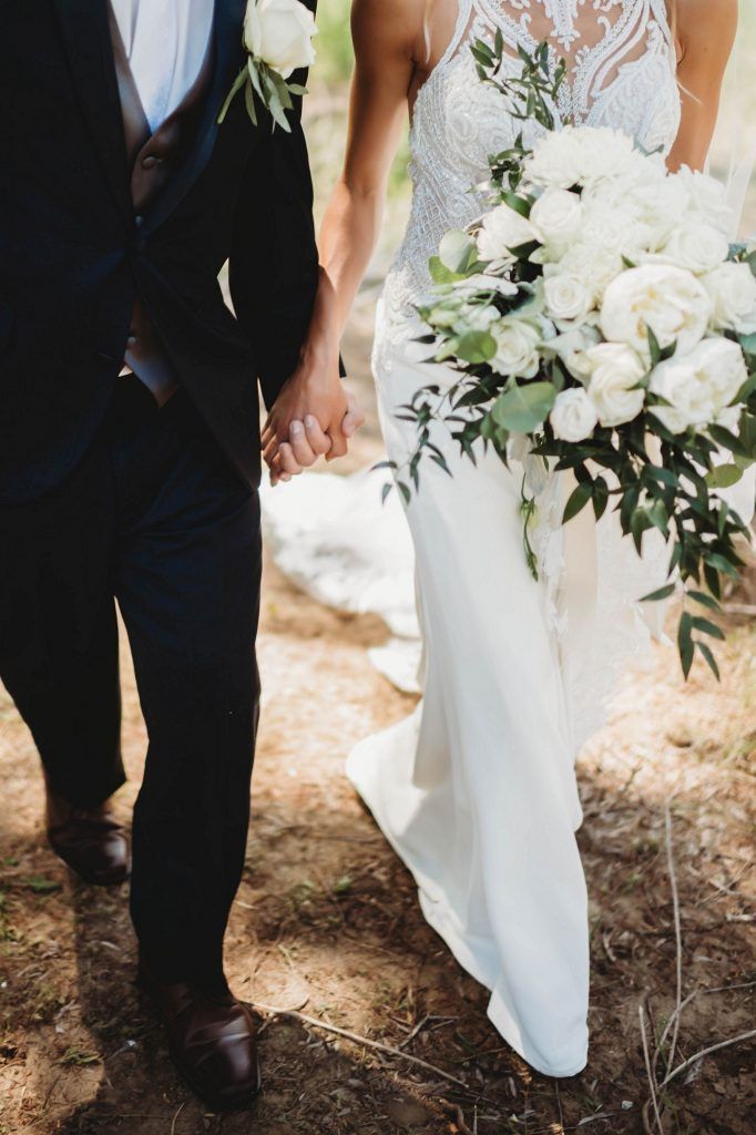 Couple holding hands, bride in white dress holding bouquet, groom in navy suit, outdoor setting.