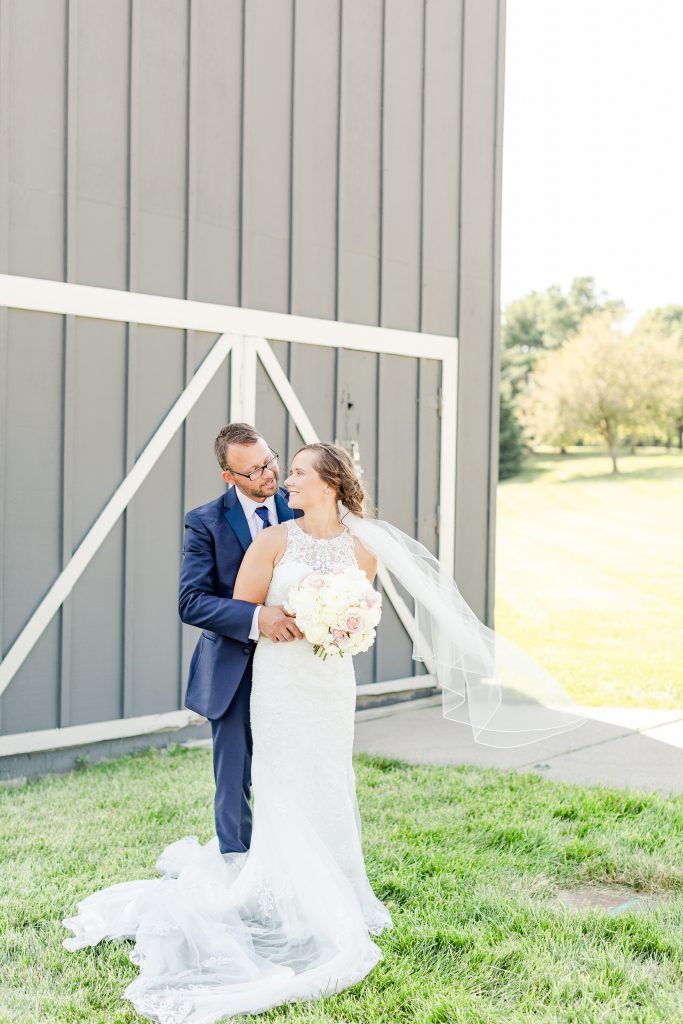 Newlyweds embracing outdoors, near a gray building. The bride wears a white dress, holding a bouquet,and the groom wears suit