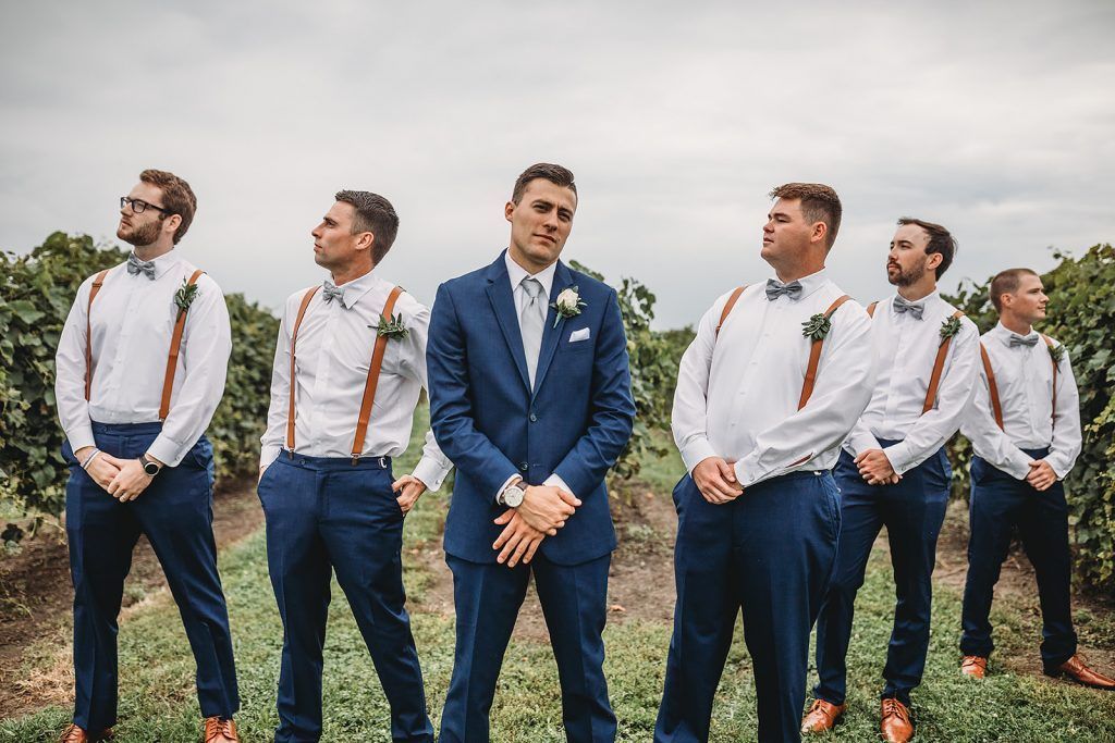 Groom and groomsmen in navy and white with suspenders, posing outdoors in a field, looking at the camera.