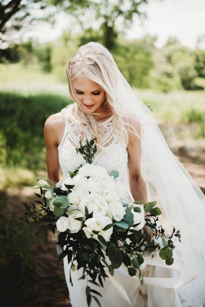 Bride in a white dress and veil holds a bouquet, walking outdoors.