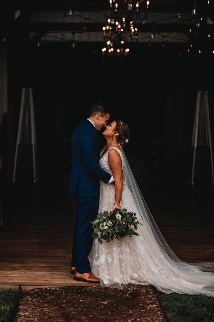 Couple kissing, bride in white dress, groom in blue suit, holding bouquet, dark backdrop.