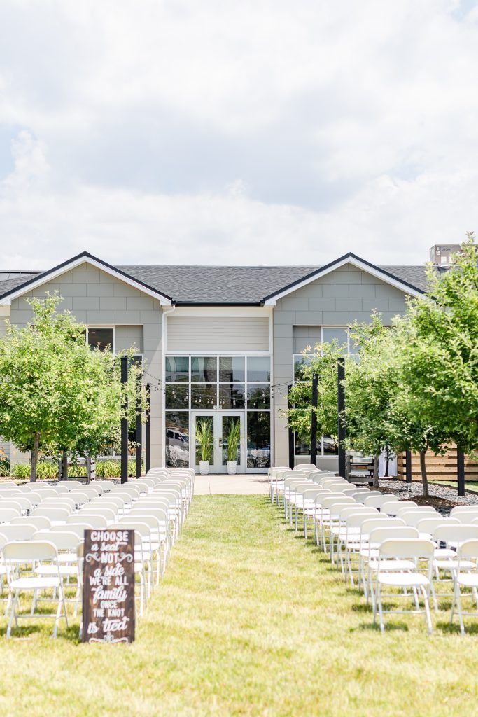 Wedding ceremony setup on a lawn; white chairs face a building with glass doors, flanked by trees and a sign.