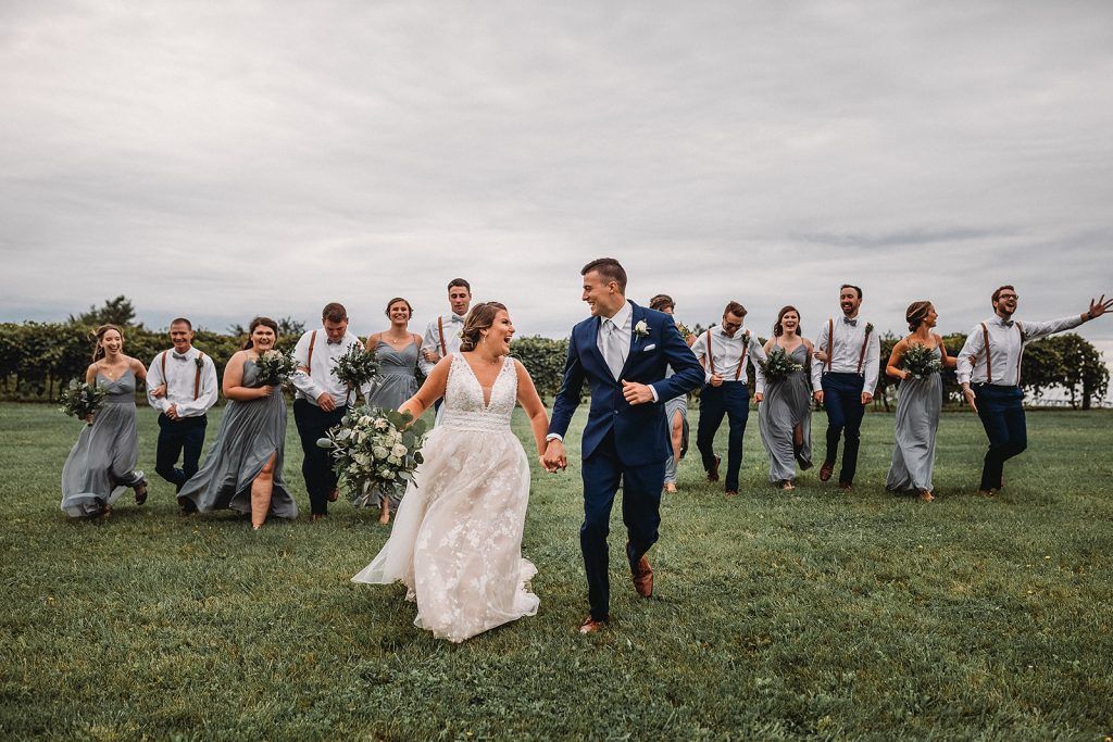 Bride and groom run, holding hands, followed by wedding party across grassy field.