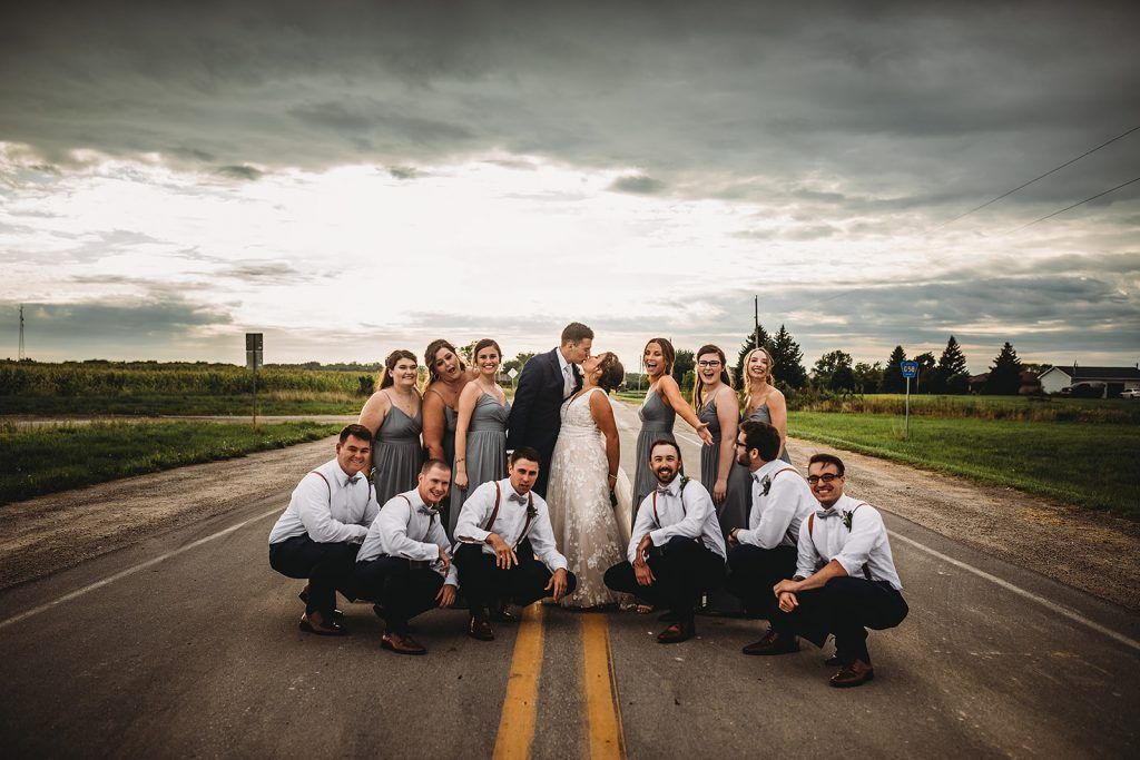 Wedding party poses on a road. Bride and groom kiss, surrounded by bridesmaids and groomsmen, under a cloudy sky.