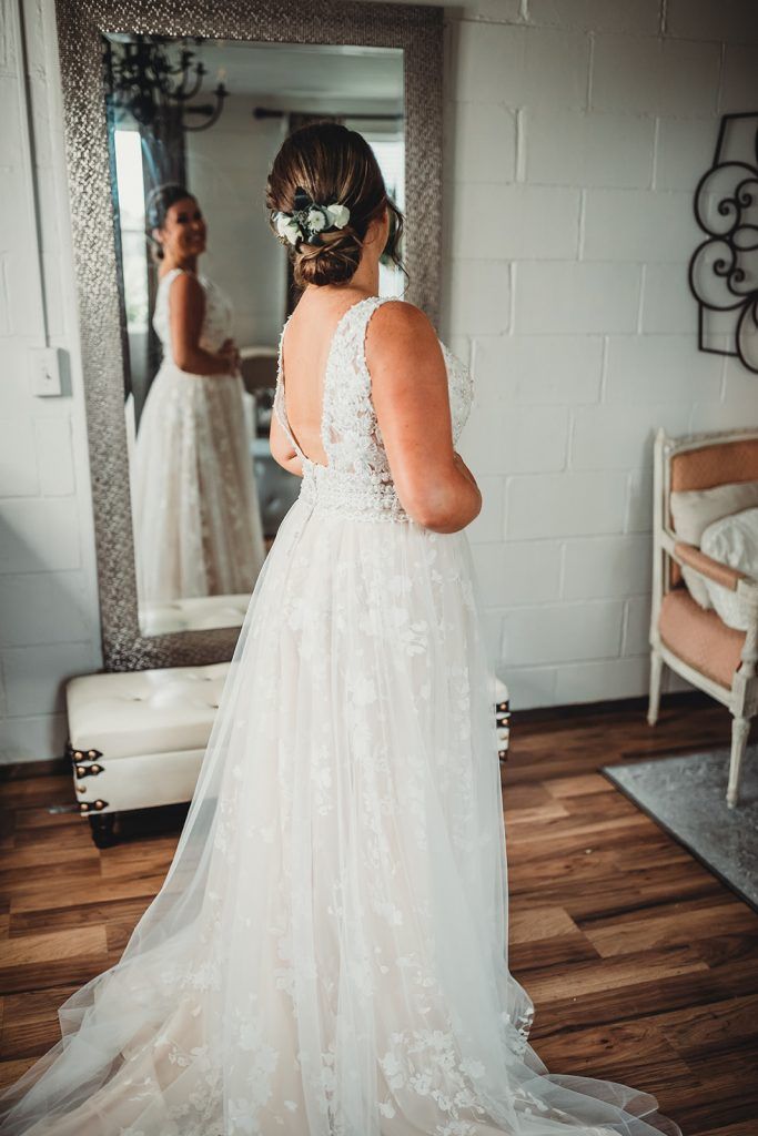 Bride in wedding dress, looking in mirror, floral hairpiece, in a room with a mirror and chair.