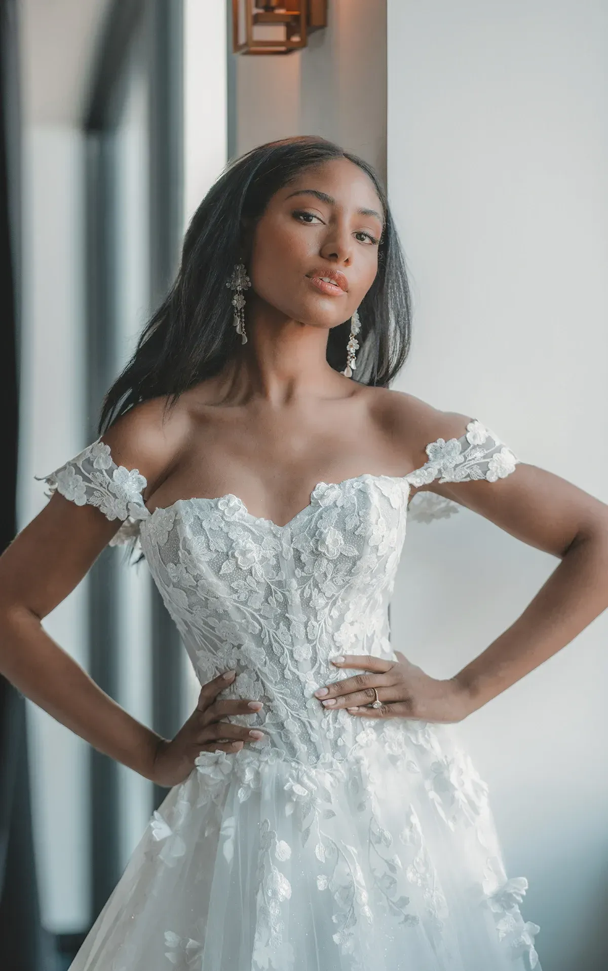 Woman in a white off-the-shoulder wedding dress, hands on hips, posing by a window.