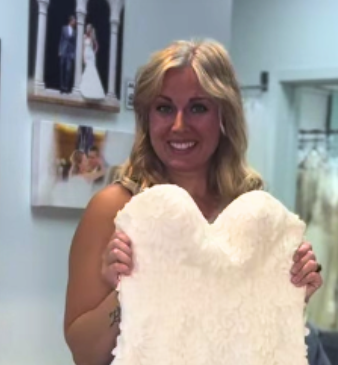 Woman holding up a white lace wedding dress, smiling inside a bridal shop.