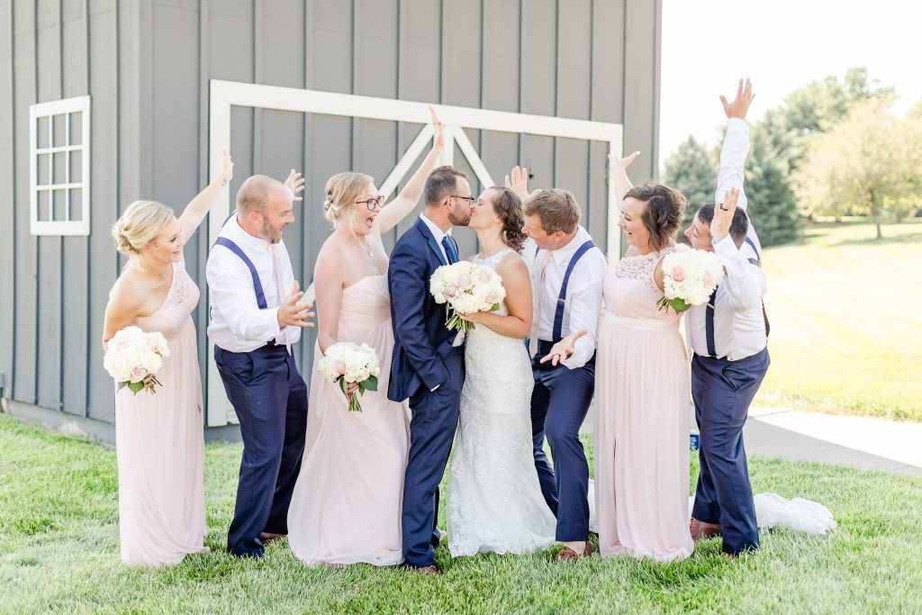 Wedding party with bride and groom kissing, raising arms near a gray barn.