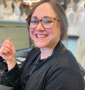Woman wearing glasses, smiling at a desk in front of a computer, with wedding dresses in the background.