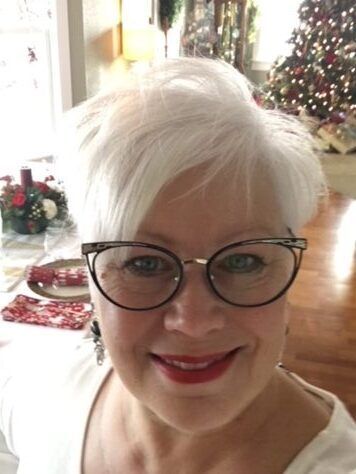 Woman with glasses smiles; Christmas tree in background.
