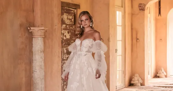Woman in a white wedding dress stands near a fountain in an outdoor setting.