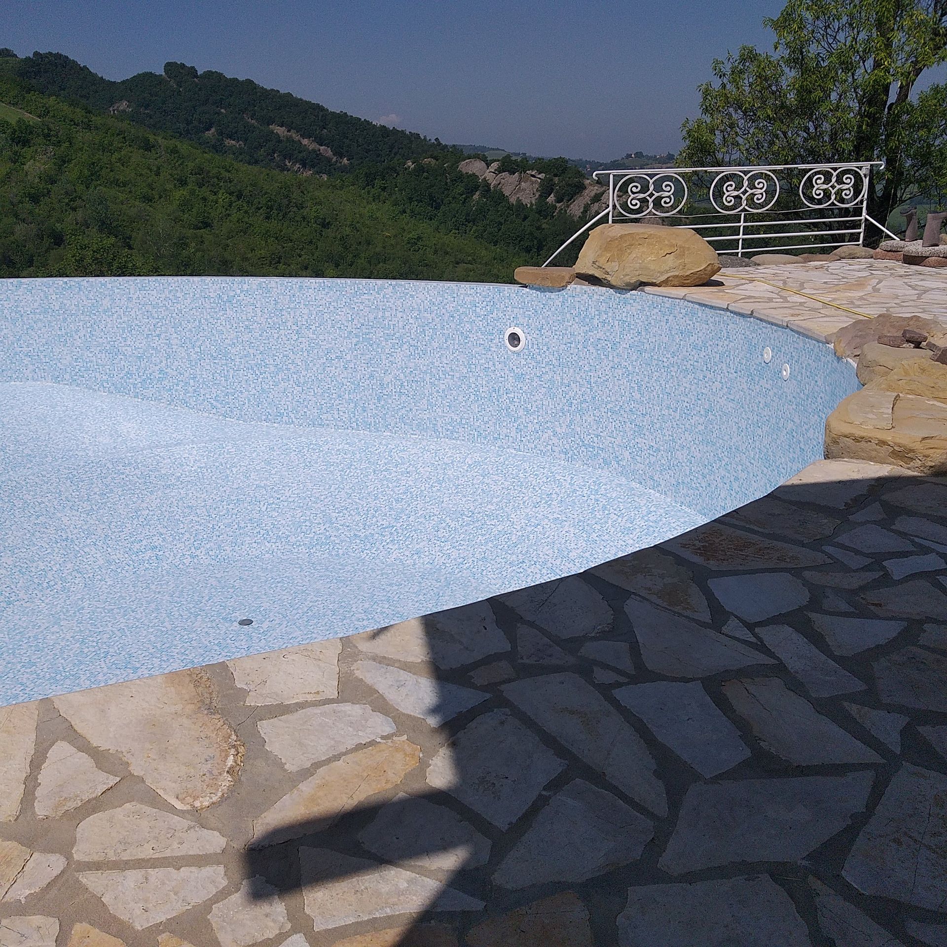 Piscina bianca vuota accanto a un patio in pietra, con verdi colline e una ringhiera metallica sullo sfondo.