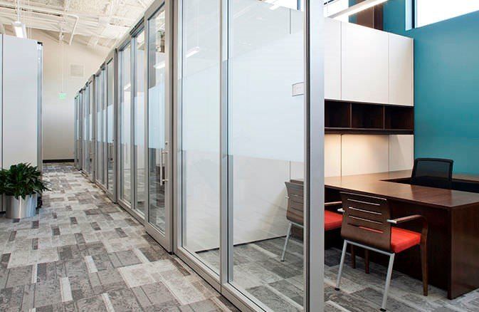 A row of office cubicles with glass walls and a desk.