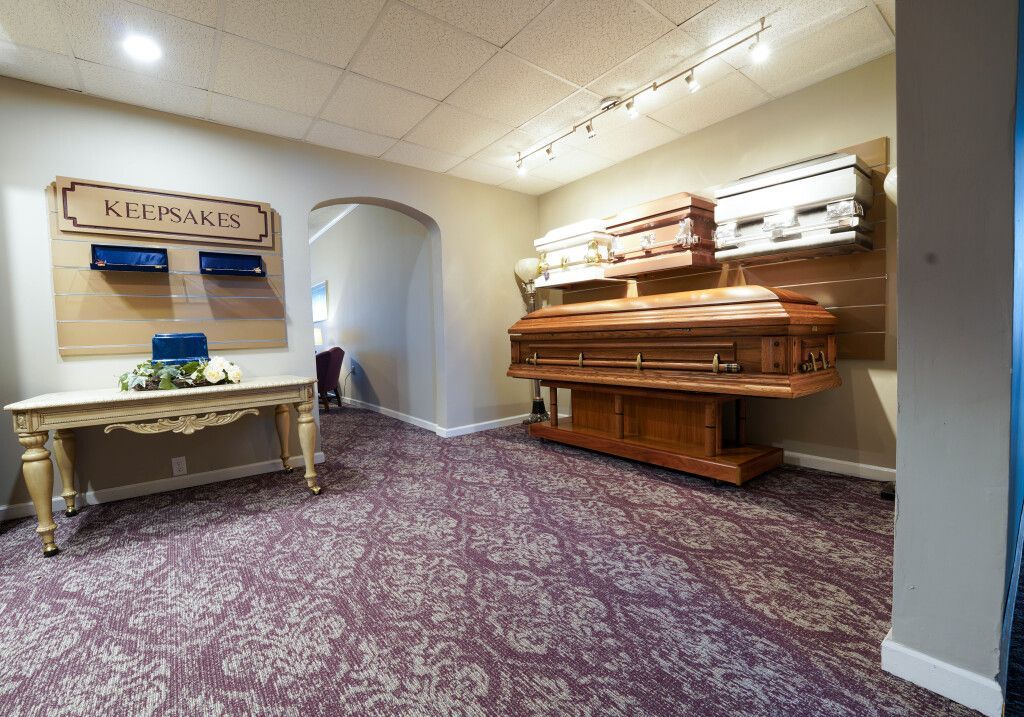 Funeral home interior. Caskets on display. Table with a floral arrangement, carpeted floor.