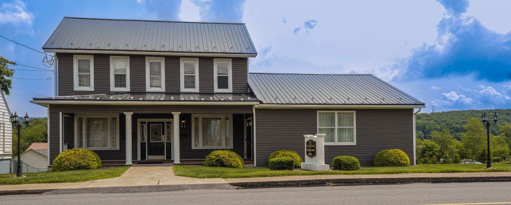 Two-story dark gray building with columns and a sloped roof, bushes in front, sky in the background.