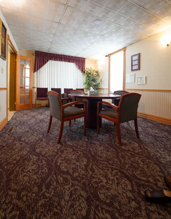 Meeting room with round table, chairs, burgundy patterned carpet, window with vertical blinds.