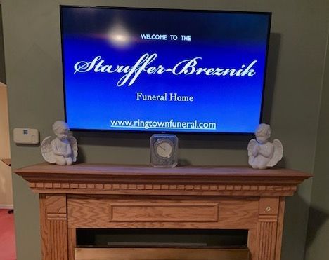 TV screen with funeral home welcome message above a fireplace mantel with angel figurines.
