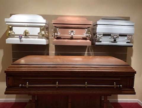 Display of caskets, including silver, copper, and wood, arranged on a wall and table in a funeral home.
