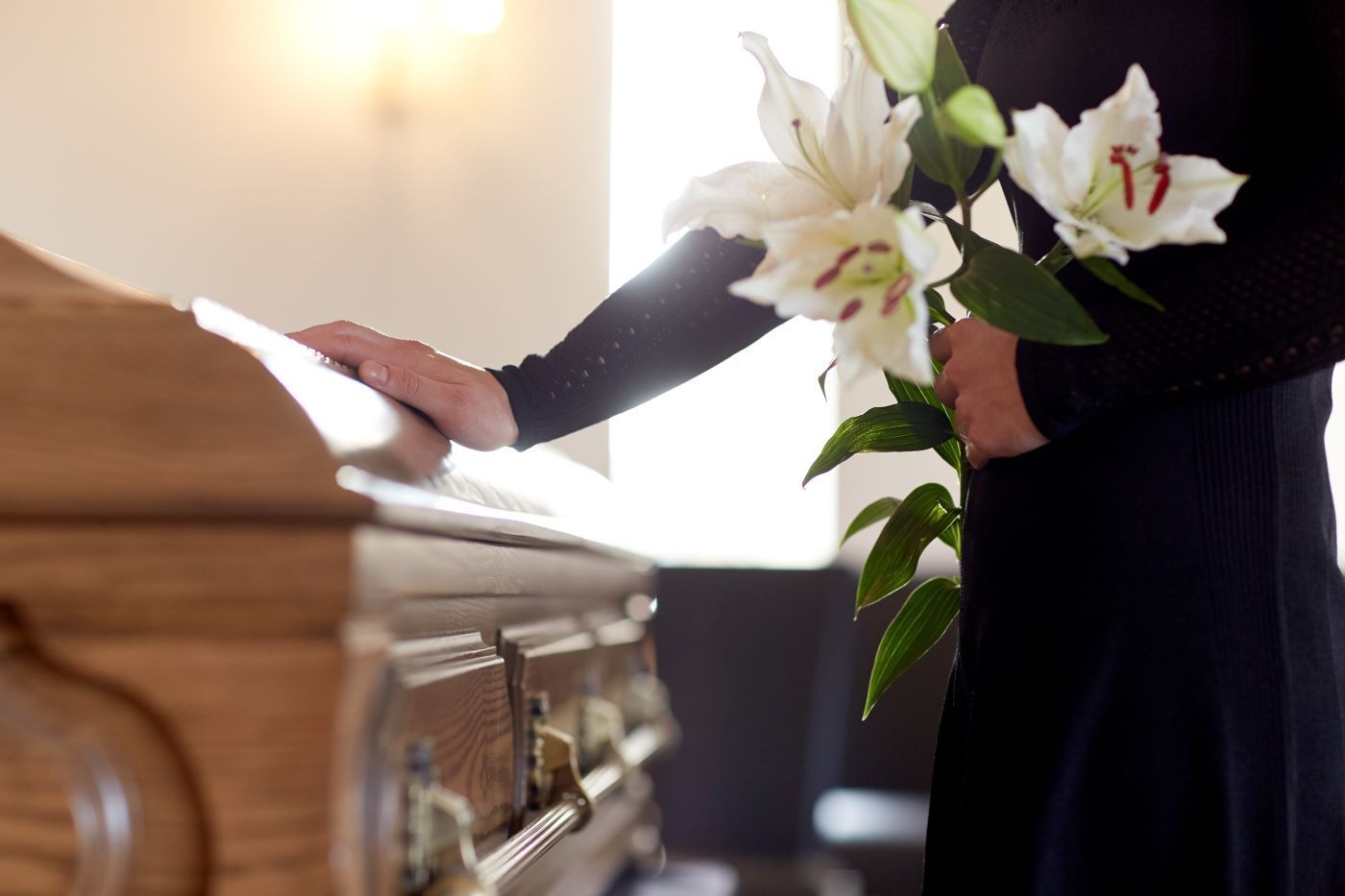 Person in black attire with a bouquet of white lilies, touching a wooden casket in a bright room.