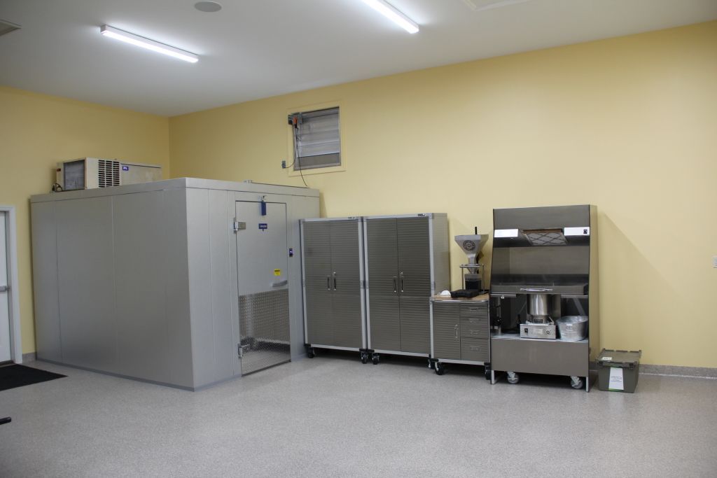 A commercial kitchen interior with stainless steel equipment, including a large freezer, cabinets, and a coffee machine, against yellow walls and a speckled floor.