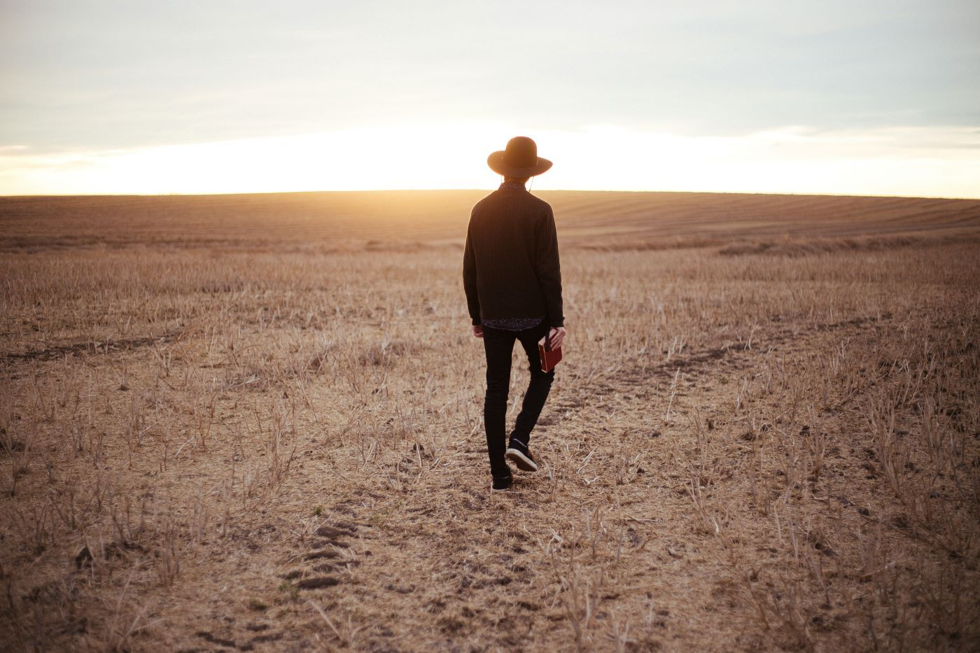 Person with a hat walks across a dry, flat field towards a sunset.