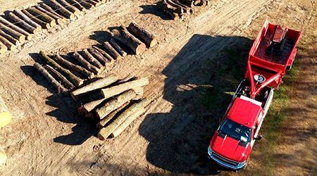 Red truck towing a trailer loaded with logs on a dirt lot.