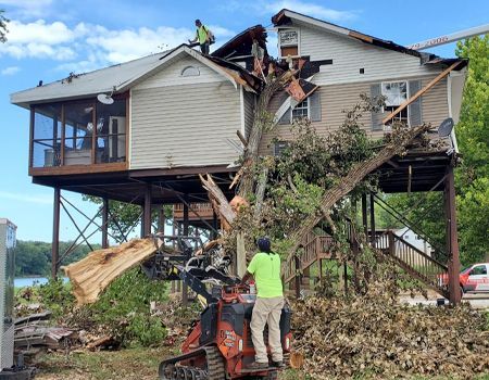 Workers removing a tree from a two-story stilt house with a destroyed roof.