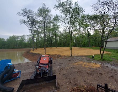 A small construction vehicle sits near a dirt-filled area with a pond on the left and trees in the background.