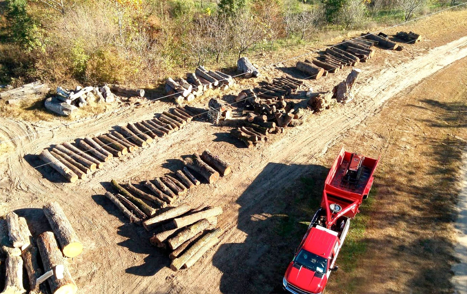Red truck with trailer next to several stacks of logs on a dirt path in a wooded area.