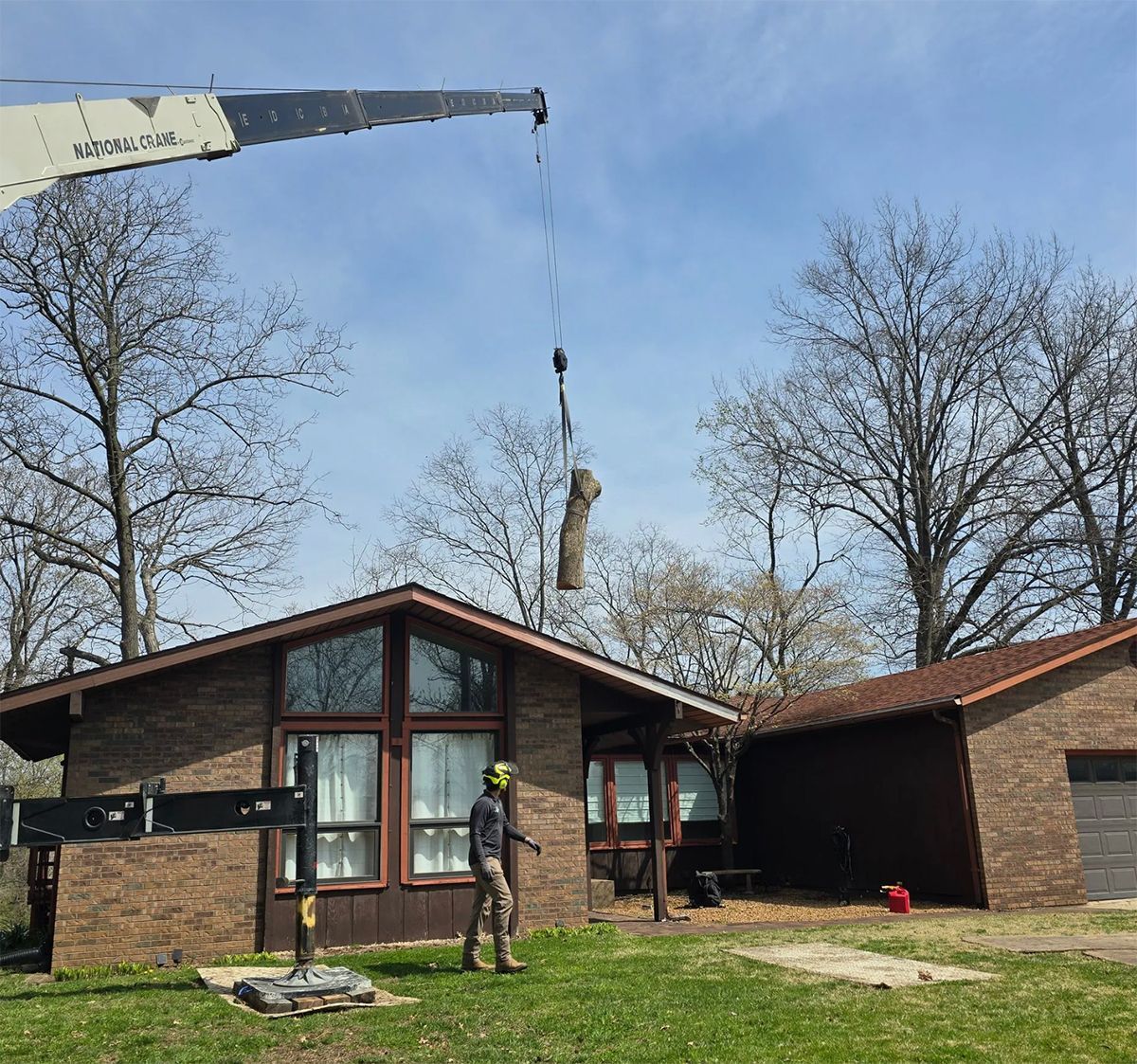 A tree trunk being lifted by a crane near a brown brick house; a worker watches on a green lawn.