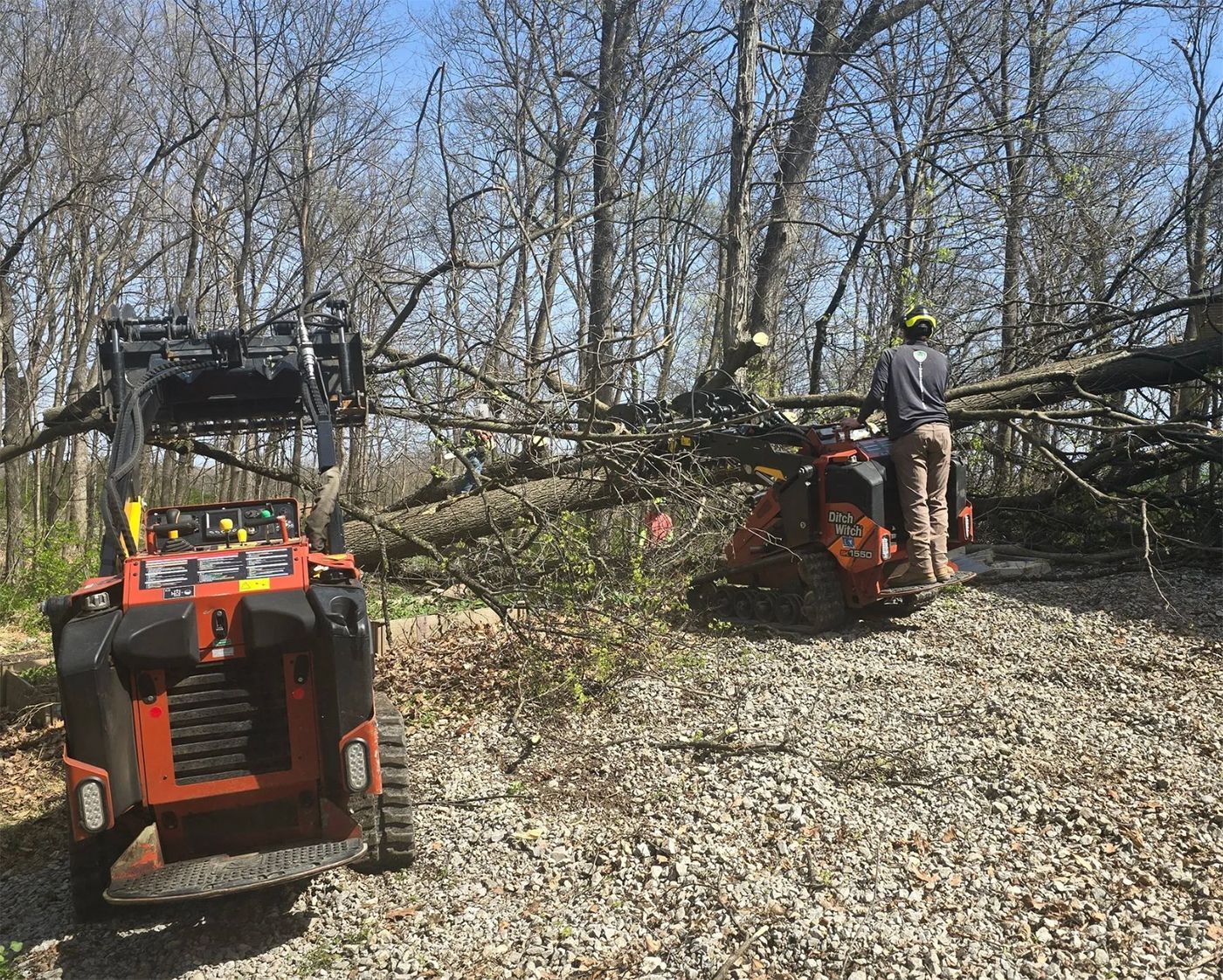 Two small orange machines clear fallen tree branches in a wooded area; a person on one operates a saw.