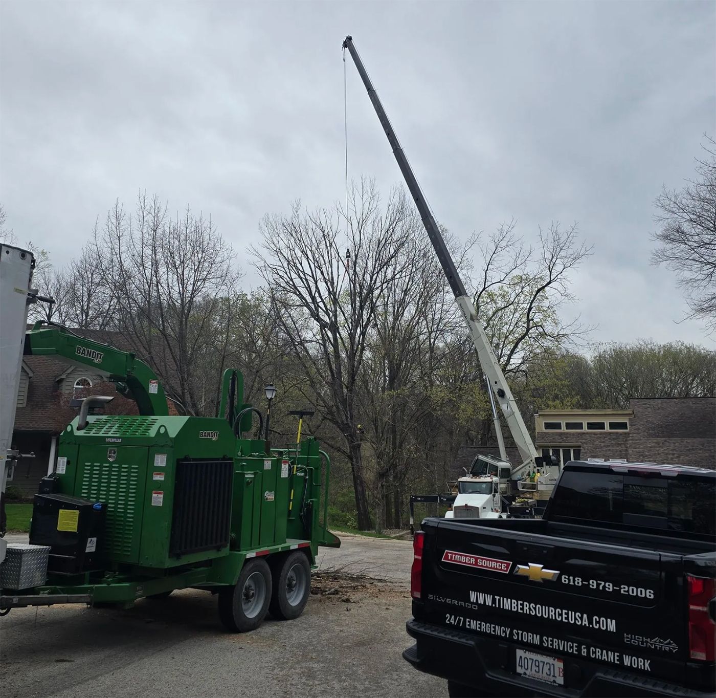 A tree removal scene with a crane, chipper, and pickup truck on a gravel driveway near a residential area.