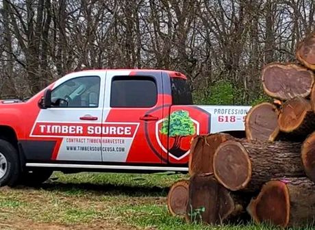 Red and white Timber Source truck next to a pile of logs outdoors.