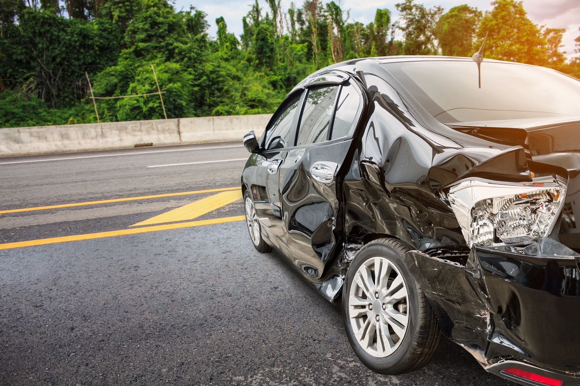 Severely damaged black car on a wet highway after a crash, with debris and trees in the background