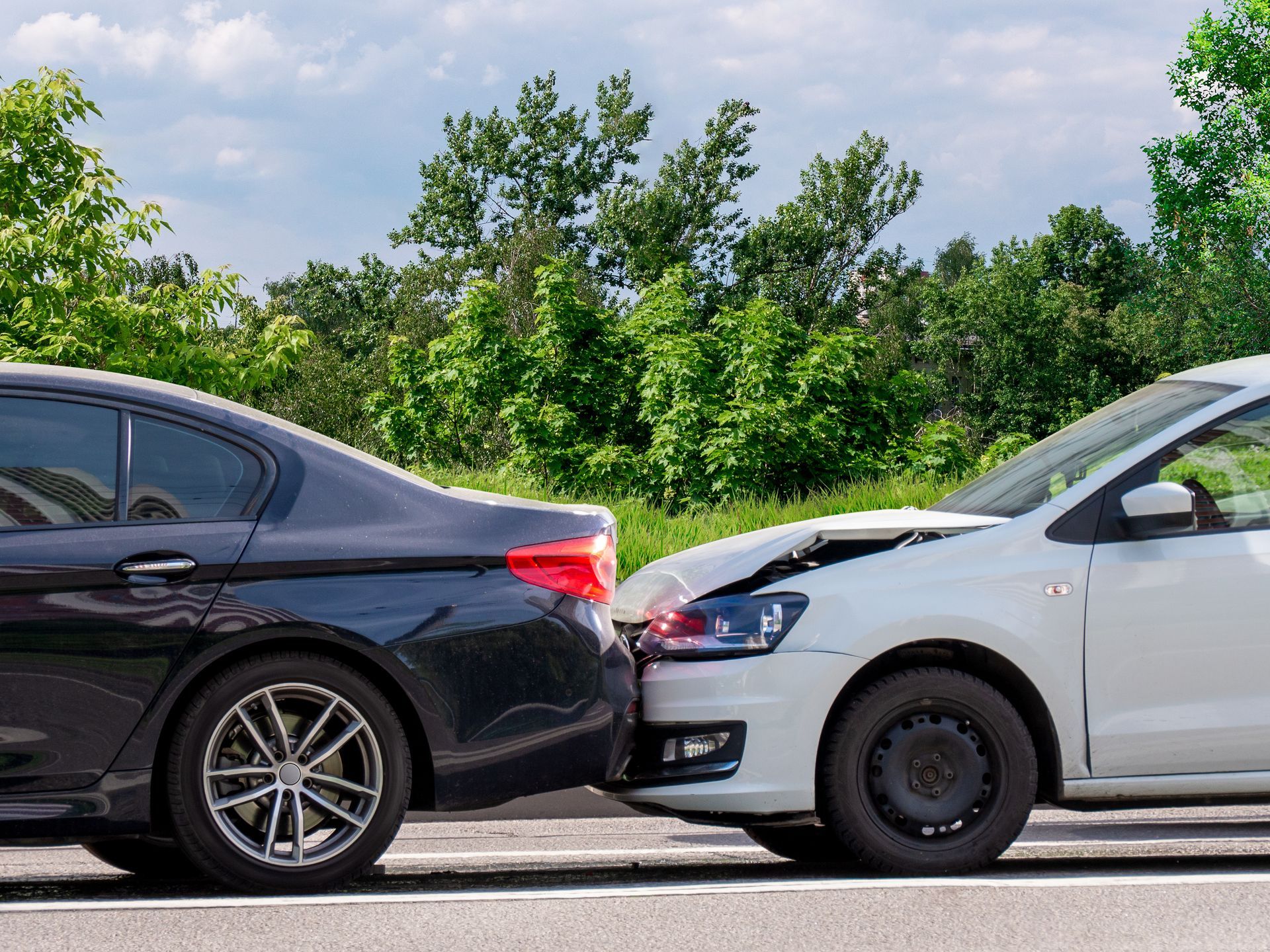 Two cars collided front-to-rear on a road beside green trees under a cloudy sky.