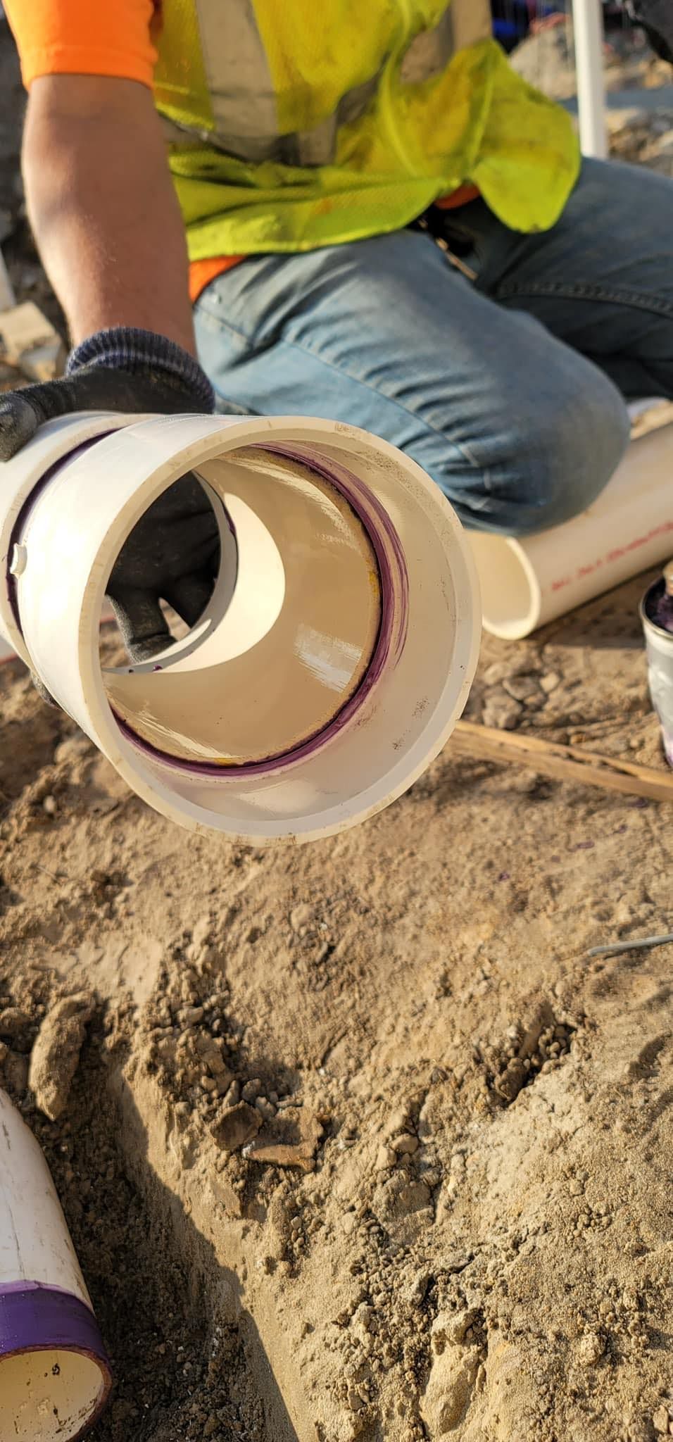 A construction worker in a high-visibility vest holds a section of white PVC pipe with a purple-primed interior edge.