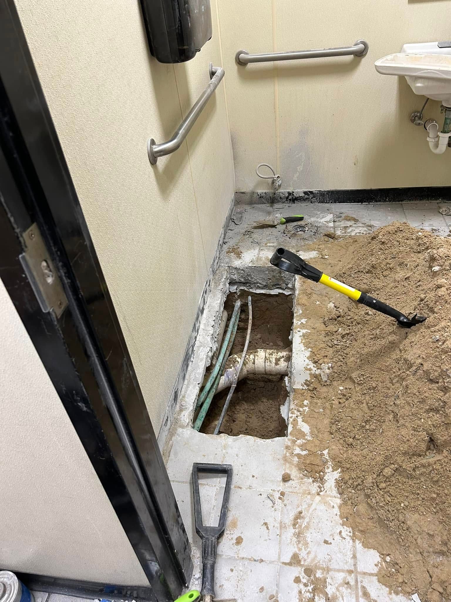 A bathroom floor with a rectangular hole exposing plumbing pipes, surrounded by construction debris and hand tools.