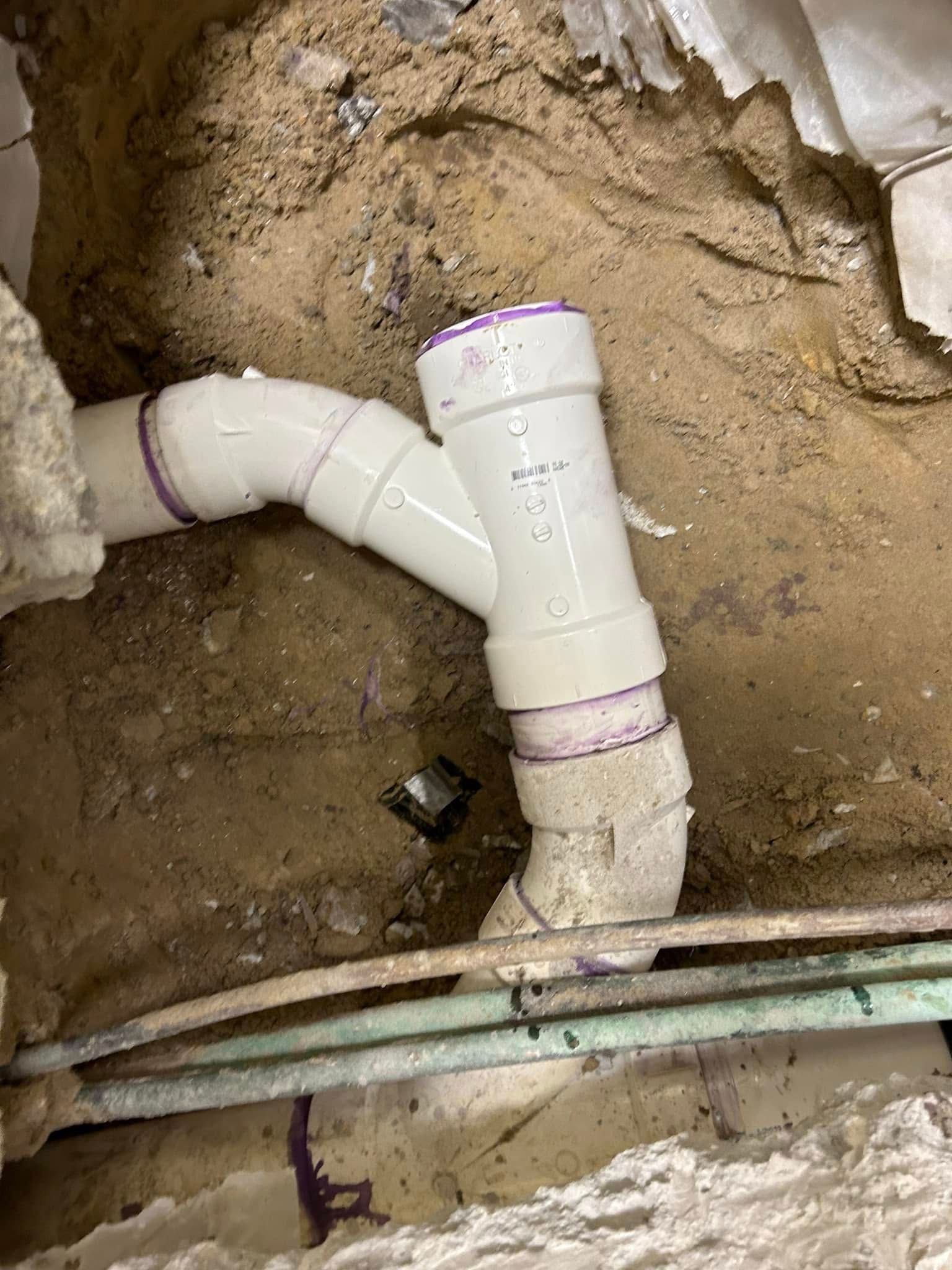 A white PVC pipe assembly with a Y-fitting and elbow installed in a dirt trench during plumbing construction.