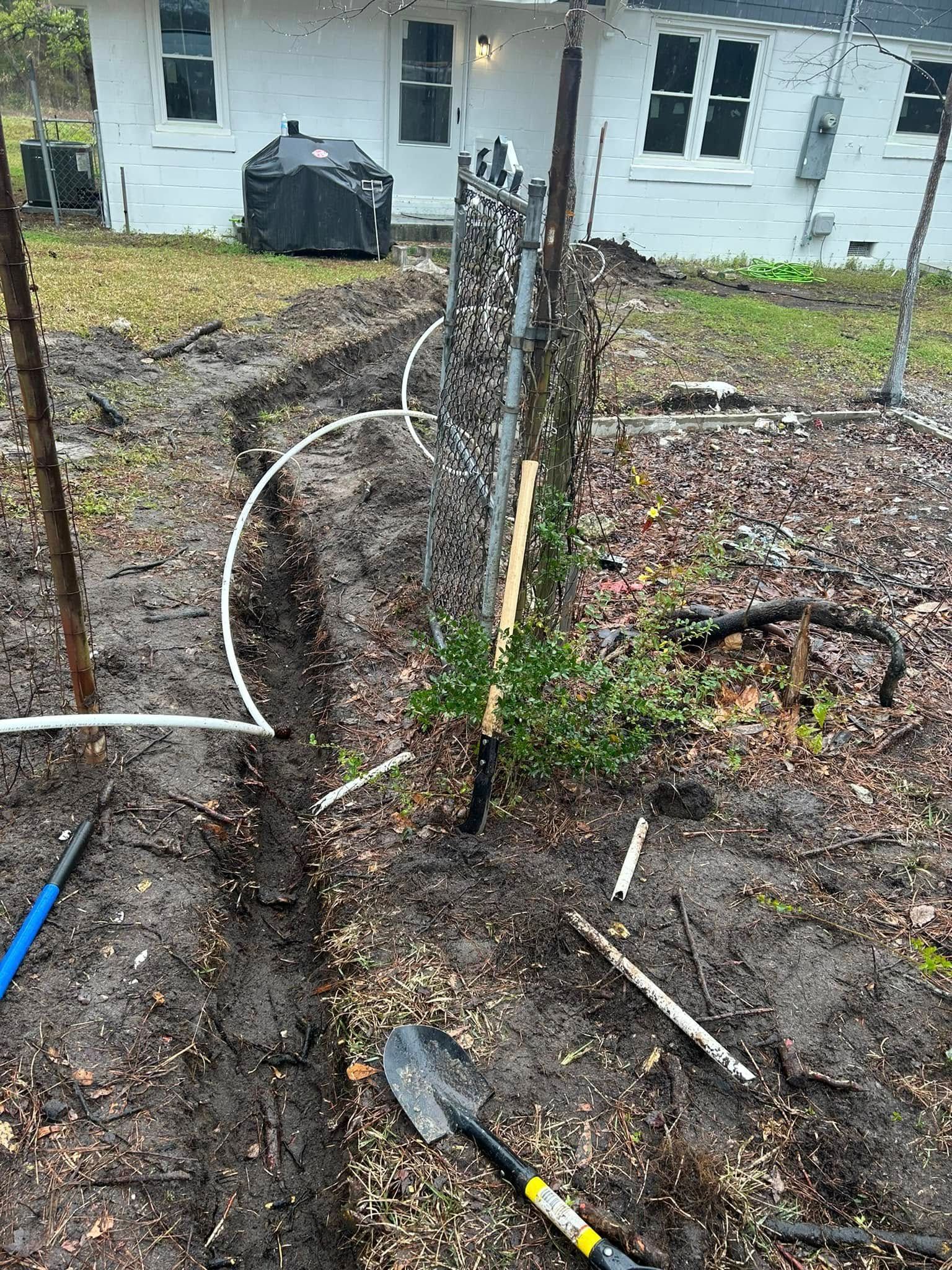 A trench dug alongside a chain-link fence in a yard with a house in the background.